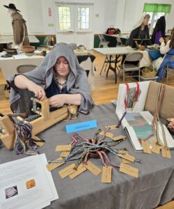 A woman wearing a gray linen veil adjusts a loom while seated at a table with various woven bands on display.